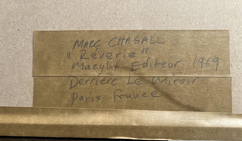 May include: A close-up of a brown paper label with handwritten text. The text reads "Marc Chagall, R&ecirc;verie, Maeght Editeur, 1969, Derriere Le Miroir, Paris France."