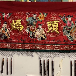 Puede incluir: Un textil rojo rectangular y largo con caracteres chinos dorados y figuras bordadas. El textil tiene un borde verde y una franja de borlas en la parte inferior. Las figuras visten ropa tradicional colorida.
