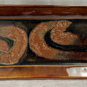 Op de afbeelding: Een rechthoekige serveerschaal van keramiek met een bruine glazuur en zwarte en koperen swirls. De schaal heeft een licht verhoogde rand.