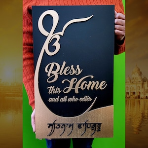 Puede incluir: Un letrero rectangular de madera negra con detalles dorados. El letrero presenta las palabras "Bless this Home and all who enter" en una elegante escritura, junto con un s&iacute;mbolo religioso y texto en otra escritura. El letrero es sostenido por una persona.