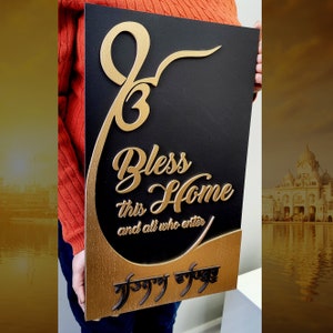 Puede incluir: Un letrero rectangular de madera negra con letras doradas. El texto dice "Bless this Home and all who enter". Presenta un s&iacute;mbolo religioso estilizado y texto en otra escritura. Los bordes del letrero son dorados.