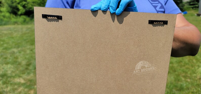 Puede incluir: Parte trasera de un panel de arte rectangular de color marr&oacute;n claro, con dos colgadores de dientes de sierra negros. El panel tiene un logotipo "ART DECORS" en la esquina inferior derecha.