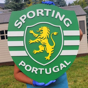 May include: A green shield-shaped sign with the words "SPORTING PORTUGAL" in white. The sign features a yellow lion emblem within a white and green crest design. The sign is held up by a person wearing blue gloves.