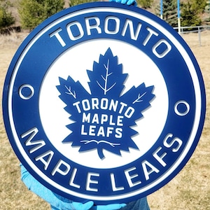 May include: A round, blue and white Toronto Maple Leafs sign. The sign features the team's logo, a stylized maple leaf, and the words "TORONTO MAPLE LEAFS" in white lettering. The sign is held up outdoors.
