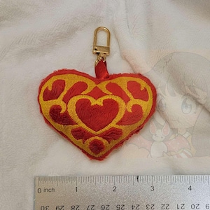 May include: A red and gold heart-shaped plush keychain with a gold metal ring. The heart has a detailed design with a red center and gold accents.
