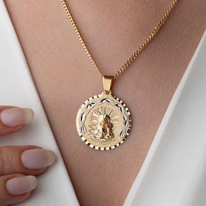 May include: A gold-toned necklace featuring a circular pendant with a central embossed figure surrounded by a sunburst pattern and a textured border. The pendant hangs from a delicate gold-toned chain, resting against a white fabric garment.