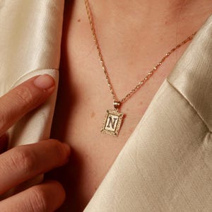 May include: A gold-toned necklace featuring a rectangular pendant with a decorative border and the letter N engraved in the center. The pendant hangs from a delicate gold-toned chain, resting against a light-colored, smooth-textured fabric garment.