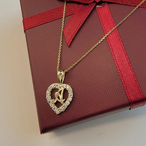 May include: A gold necklace featuring a heart-shaped pendant with the letter "A" and clear crystals. The necklace is displayed on a burgundy gift box tied with a red ribbon.