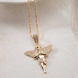 May include: A gold-toned necklace featuring a pendant shaped like a cherub with wings and hands pressed together in prayer. The cherub's wings and the pendant's bail are encrusted with small, clear stones. The pendant hangs from a gold-toned rope chain.