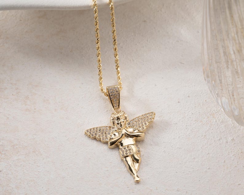 May include: A gold-toned necklace featuring a pendant shaped like a cherub with wings and hands pressed together in prayer. The cherub's wings and the pendant's bail are encrusted with small, clear stones. The pendant hangs from a gold-toned rope chain.