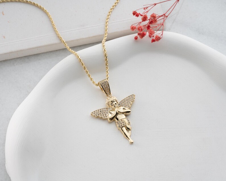May include: A gold-toned necklace featuring a pendant shaped like a cherub with outstretched wings. The pendant is embellished with small, clear, sparkling stones covering the wings and the bail. The chain is a twisted rope style in gold metal.