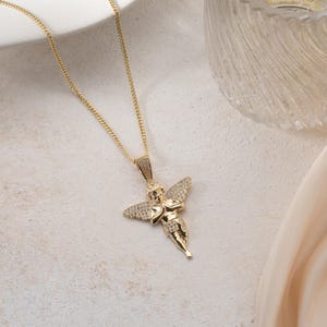 May include: A gold-toned necklace featuring a pendant shaped like a cherub with outstretched wings. The pendant is embellished with small, clear, sparkling stones covering the wings and the figure's body, suspended from a delicate gold-toned chain.