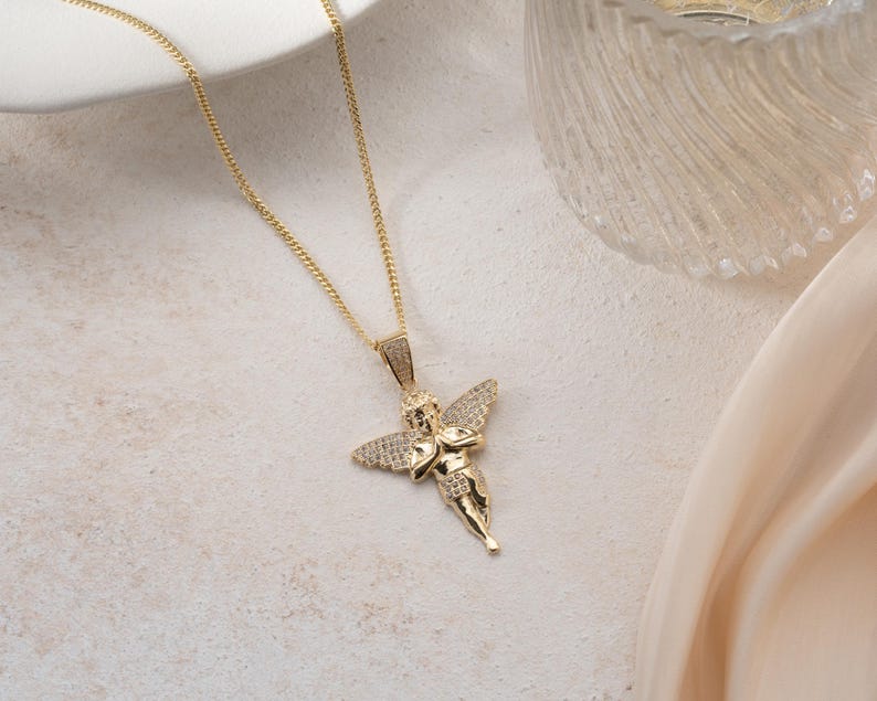 May include: A gold-toned necklace featuring a pendant shaped like a cherub with outstretched wings. The pendant is embellished with small, clear, sparkling stones covering the wings and the figure's body, suspended from a delicate gold-toned chain.