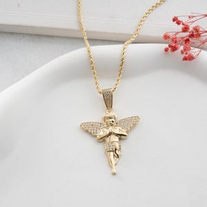 May include: A gold-toned necklace featuring a pendant shaped like a cherub with wings, adorned with small clear stones. The cherub is depicted with hands pressed together in a prayer position. The pendant hangs from a twisted gold-toned rope chain.