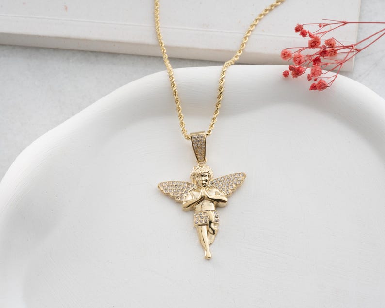 May include: A gold-toned necklace featuring a pendant shaped like a cherub with wings, adorned with small clear stones. The cherub is depicted with hands pressed together in a prayer position. The pendant hangs from a twisted gold-toned rope chain.