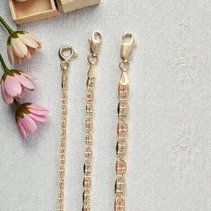 Op de afbeelding: Drie goudkleurige kettingen met verschillende kettingontwerpen worden gepresenteerd tegen een lichtgrijze achtergrond. De kettingen hebben verschillende patronen, waaronder een delicate ketting en kettingen met geometrische vormen. Roze en witte bloemen staan op de achtergrond.
