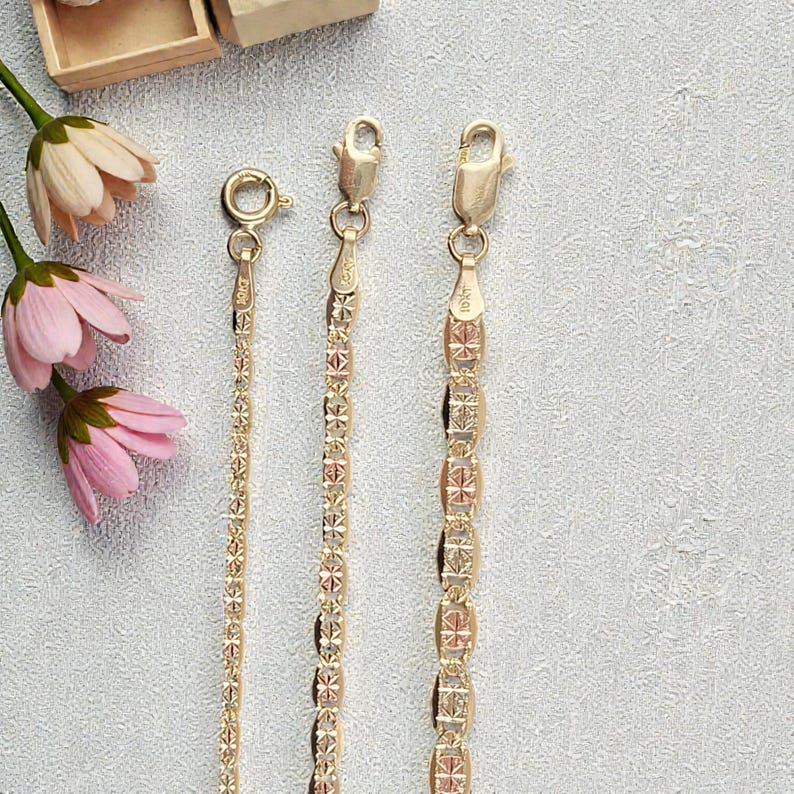 Op de afbeelding: Drie goudkleurige kettingen met verschillende kettingontwerpen worden gepresenteerd tegen een lichtgrijze achtergrond. De kettingen hebben verschillende patronen, waaronder een delicate ketting en kettingen met geometrische vormen. Roze en witte bloemen staan op de achtergrond.