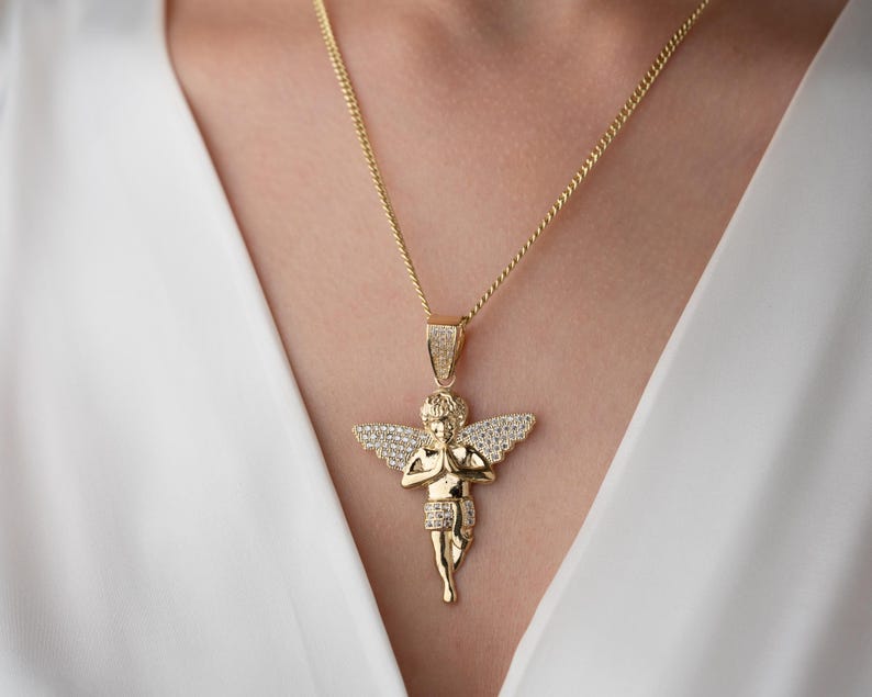 May include: A gold-toned necklace featuring a pendant shaped like a cherub with wings outstretched and hands pressed together in prayer. The cherub's wings and the pendant's bail are encrusted with small, clear, sparkling stones, while the chain is a link style.