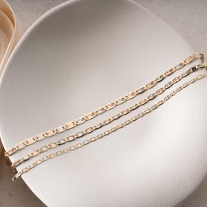 Puede incluir: Tres pulseras de cadena en tono dorado con un dise&ntilde;o de eslabones planos entrelazados. Cada pulsera muestra un acabado pulido con segmentos lisos y texturizados alternos, creando una superficie reflectante. Las cadenas est&aacute;n dispuestas en paralelo.