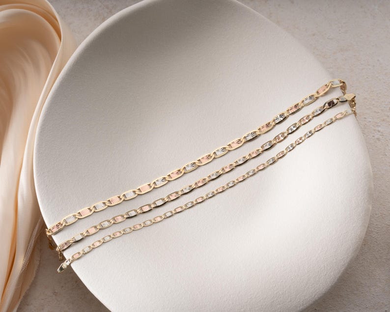 Puede incluir: Tres pulseras de cadena en tono dorado con un dise&ntilde;o de eslabones planos entrelazados. Cada pulsera muestra un acabado pulido con segmentos lisos y texturizados alternos, creando una superficie reflectante. Las cadenas est&aacute;n dispuestas en paralelo.