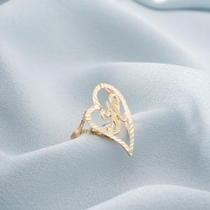 Op de afbeelding: Een goudkleurige ring met een hartvormig ontwerp. De ring heeft een open hart met een sierlijke letter "H" in het midden. De ring is geplaatst tegen een lichtblauwe, getextureerde achtergrond.