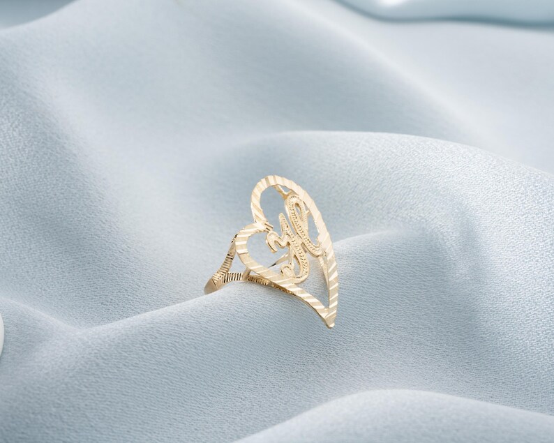 Op de afbeelding: Een goudkleurige ring met een hartvormig ontwerp. De ring heeft een open hart met een sierlijke letter "H" in het midden. De ring is geplaatst tegen een lichtblauwe, getextureerde achtergrond.