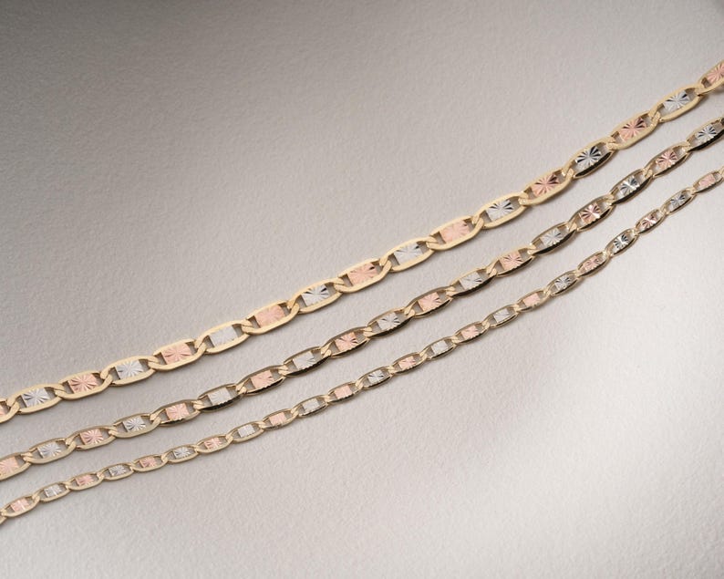 Puede incluir: Tres cadenas de oro de diferentes grosores dispuestas en diagonal. Cada cadena presenta un patr&oacute;n repetitivo de eslabones ovalados en tonos oro amarillo, blanco y rosa, con superficies texturizadas y facetadas que reflejan la luz sobre los eslabones.