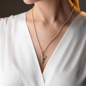May include: A gold-toned rope chain necklace featuring a pendant shaped like a cherub with outstretched wings. The angel pendant is embellished with small, clear, sparkling stones set across the wings and body, creating a reflective surface.