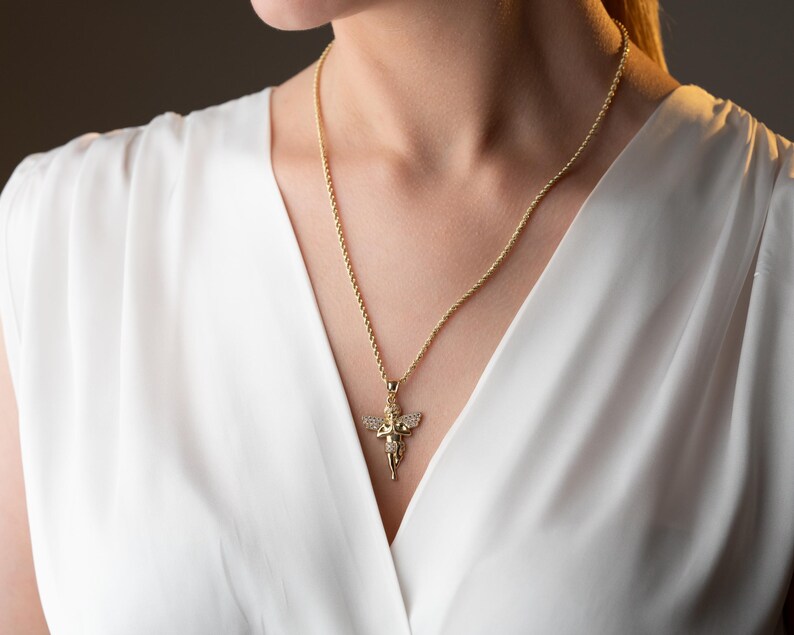 May include: A gold-toned rope chain necklace featuring a pendant shaped like a cherub with outstretched wings. The angel pendant is embellished with small, clear, sparkling stones set across the wings and body, creating a reflective surface.