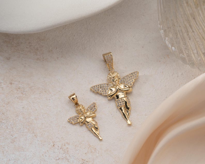 May include: Two gold-toned angel pendants featuring pav&eacute;-set clear stones on the wings and lower garments. Each pendant depicts a cherub figure with hands clasped in a prayer position, suspended from a small integrated bail at the top of the head.