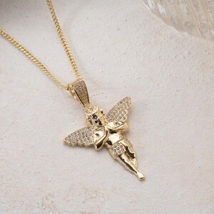 May include: A gold-toned necklace featuring a pendant shaped like a cherub with outstretched wings. The pendant is embellished with small, clear pav&eacute;-set stones covering the wings and the bail, while the figure itself has a polished gold finish.