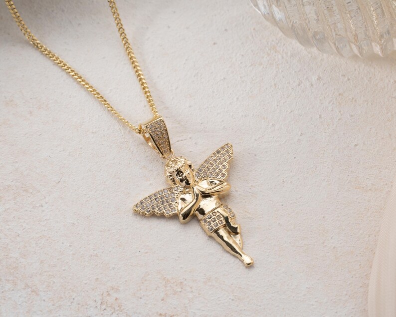 May include: A gold-toned necklace featuring a pendant shaped like a cherub with outstretched wings. The pendant is embellished with small, clear pav&eacute;-set stones covering the wings and the bail, while the figure itself has a polished gold finish.