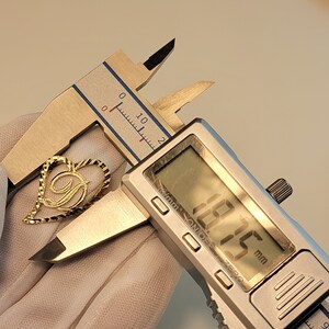 Op de afbeelding: Een goudkleurige hartvormige hanger met de letter "D" erin, gemeten met een digitale schuifmaat. De schuifmaat geeft een meting van 18,75 mm weer. De hanger heeft een getextureerde rand.