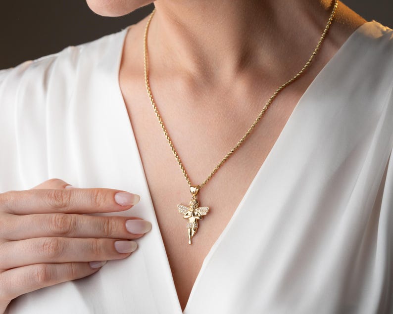 May include: A gold-toned rope chain necklace featuring a small, detailed angel pendant. The angel figure has outstretched wings and is embellished with small, clear, sparkling stones across its surface, suspended from a simple gold-toned bail.