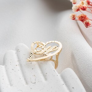Op de afbeelding: Een goudkleurige ring in de vorm van een hart, met de letter "M" in het midden. De ring wordt gepresenteerd op een wit oppervlak, met een vage achtergrond en een paar bloesemtakjes.