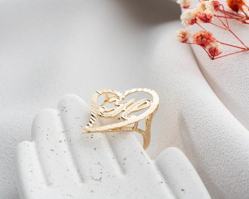 Op de afbeelding: Een goudkleurige ring in de vorm van een hart, met de letter "M" in het midden. De ring wordt gepresenteerd op een wit oppervlak, met een vage achtergrond en een paar bloesemtakjes.