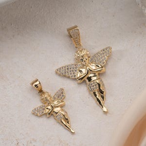 May include: Two gold-toned angel pendants of different sizes, each featuring a cherub figure with hands clasped in prayer. The wings and lower garments of both pendants are encrusted with small, clear, sparkling stones, and each has a bail at the top.