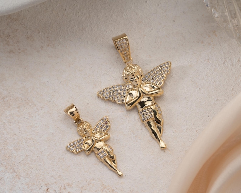 May include: Two gold-toned angel pendants of different sizes, each featuring a cherub figure with hands clasped in prayer. The wings and lower garments of both pendants are encrusted with small, clear, sparkling stones, and each has a bail at the top.