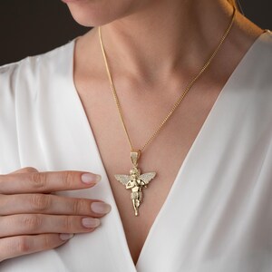 May include: A gold-toned pendant necklace featuring a cherub figure with wings, depicted in a prayer pose. The pendant is adorned with small, clear, sparkling stones embedded across the wings and body, suspended from a matching gold-toned chain.