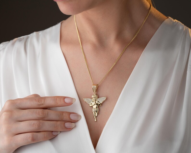 May include: A gold-toned pendant necklace featuring a cherub figure with wings, depicted in a prayer pose. The pendant is adorned with small, clear, sparkling stones embedded across the wings and body, suspended from a matching gold-toned chain.