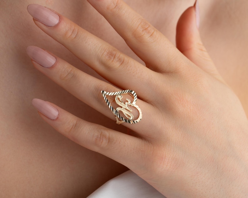 Op de afbeelding: Een goudkleurige ring met een open hartontwerp, met de letter "S" in een scriptlettertype. De ring wordt aan een vinger gedragen en toont zijn ingewikkelde details en gepolijste afwerking. Het ontwerp van de ring is een hartvorm met een gedraaide touwrand.