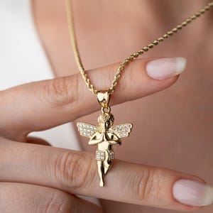 May include: A gold-toned pendant necklace featuring a cherub figure with hands pressed together in prayer. The cherub has wings and a lower garment embellished with small, clear, sparkling stones. The pendant hangs from a twisted rope-style chain.