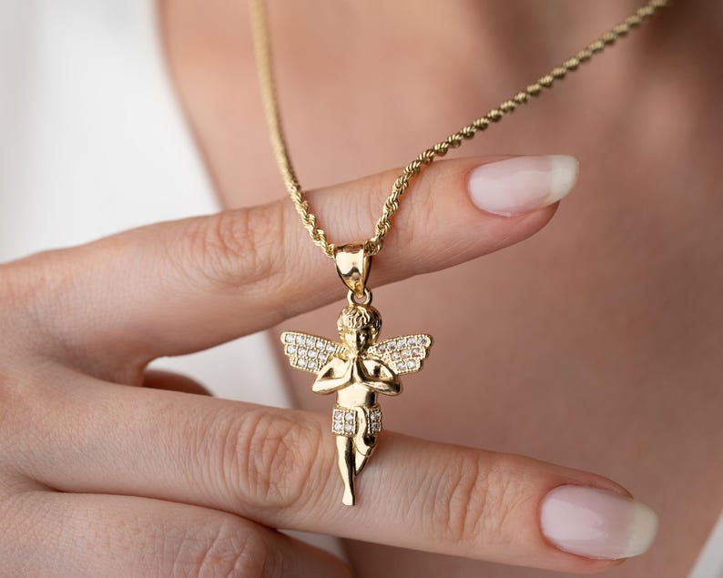 May include: A gold-toned pendant necklace featuring a cherub figure with hands pressed together in prayer. The cherub has wings and a lower garment embellished with small, clear, sparkling stones. The pendant hangs from a twisted rope-style chain.