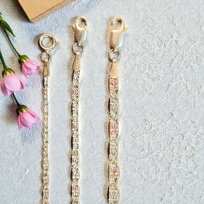 Op de afbeelding: Drie gouden armbanden met verschillende kettingontwerpen. De armbanden hebben karabijnsluitingen en worden gepresenteerd op een lichte achtergrond. Roze bloemen zijn linksboven te zien. De armbanden zijn gemarkeerd met "10KT".