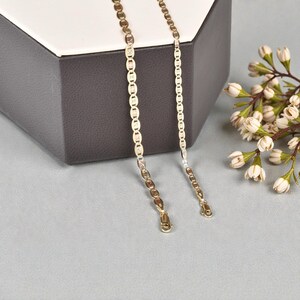 Op de afbeelding: Twee goudkleurige kettingen met verschillende kettingontwerpen. E&eacute;n heeft platte, ovale schakels, de andere een meer traditionele ketting. De kettingen worden gepresenteerd op een grijze ondergrond met een decoratieve doos en witte bloemen.