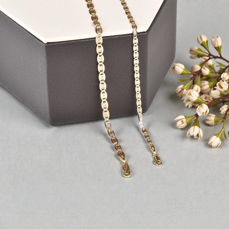 Op de afbeelding: Twee goudkleurige kettingen met verschillende kettingontwerpen. E&eacute;n heeft platte, ovale schakels, de andere een meer traditionele ketting. De kettingen worden gepresenteerd op een grijze ondergrond met een decoratieve doos en witte bloemen.