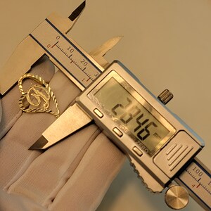 Op de afbeelding: Een goudkleurige hanger in de vorm van een hart met de letter "B" erin. Een digitale schuifmaat meet de hanger op 28,46 mm. De hanger wordt vastgehouden door een gehandschoende hand.