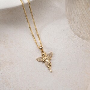 May include: A gold-toned necklace featuring a pendant shaped like a cherub with outstretched wings. The cherub figure is embellished with small, clear, sparkling stones set into the wings and body, suspended from a delicate gold-toned chain.