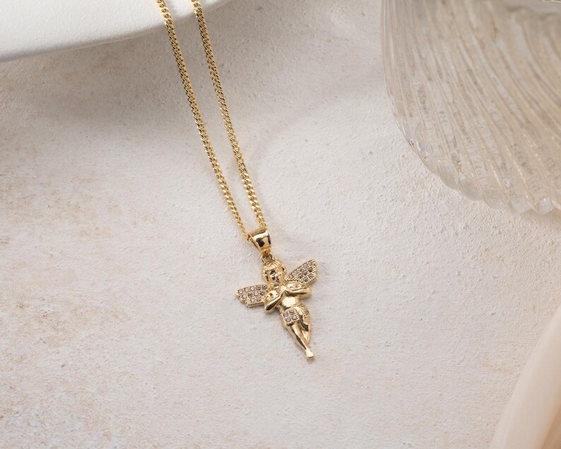 May include: A gold-toned necklace featuring a pendant shaped like a cherub with outstretched wings. The cherub figure is embellished with small, clear, sparkling stones set into the wings and body, suspended from a delicate gold-toned chain.