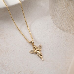 May include: A gold-toned rope chain necklace featuring a pendant shaped like a cherub with outstretched wings. The cherub figure is embellished with small, clear, sparkling stones set along the wings and body, creating a detailed metallic accessory.